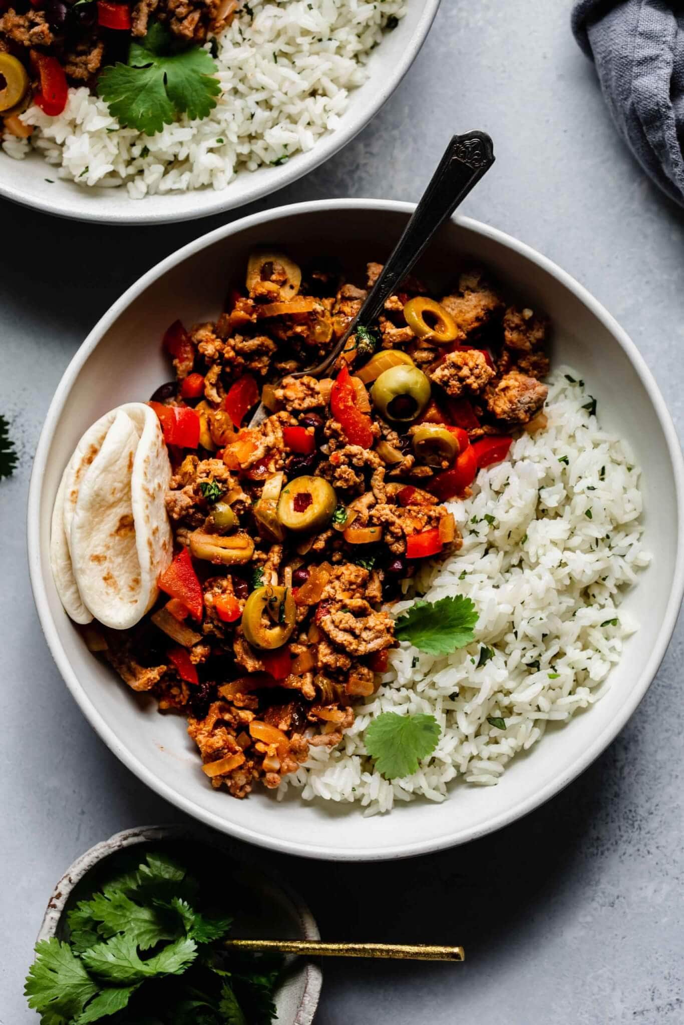 Picadillo in shallow white bowl with folded tortilla and rice.