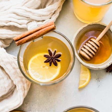 THREE MUGS OF HOT TODDIES GARNISHED WITH LEMON SLICES AND STAR ANISE NEXT TO HONEY JAR