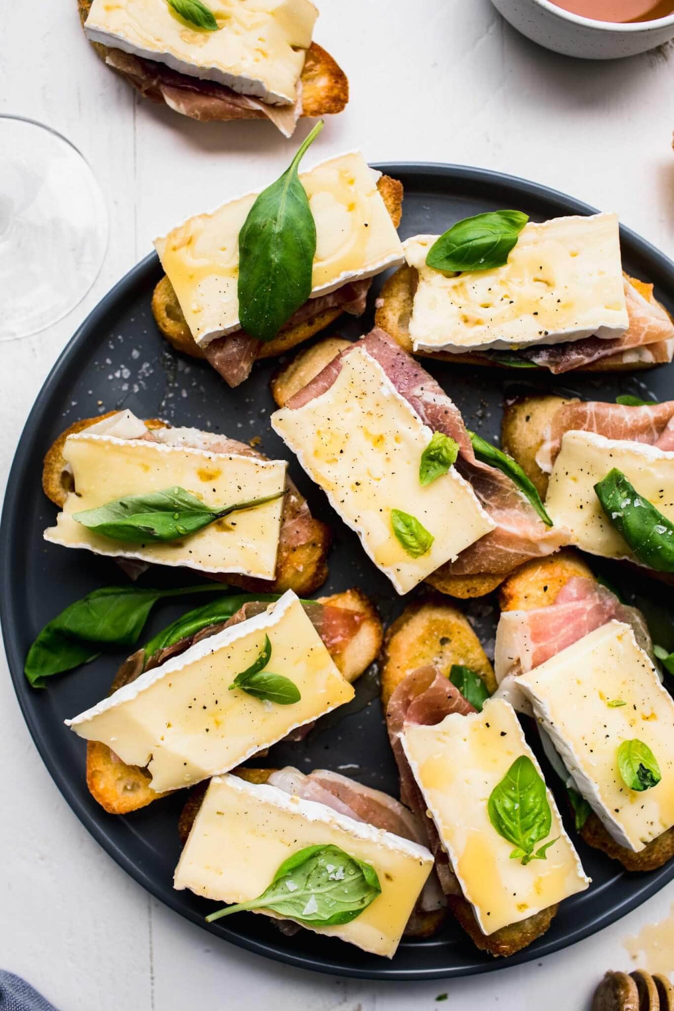 Crostini topped with brie, basil and prosciutto arranged on grey plate next to honey wand. 