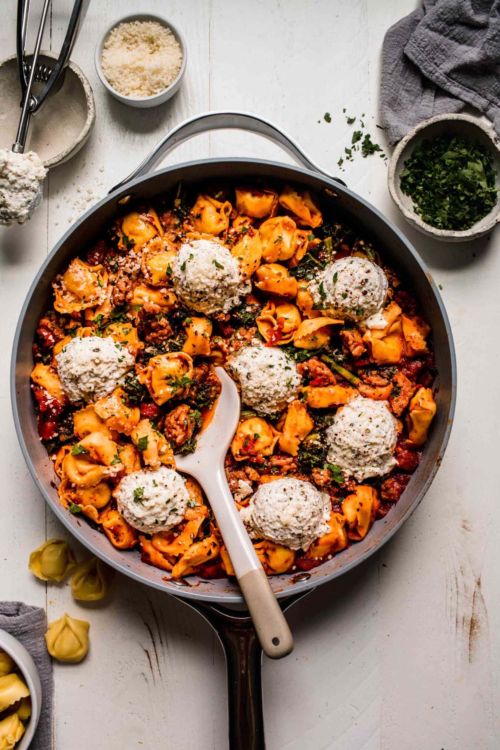 Tortellini lasagna in skillet with serving spoon next to bowls of parsley and parmesan.