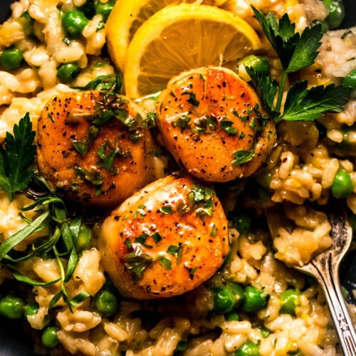 Overhead shot of scallop and pea risotto in black bowl topped with lemon wedges.