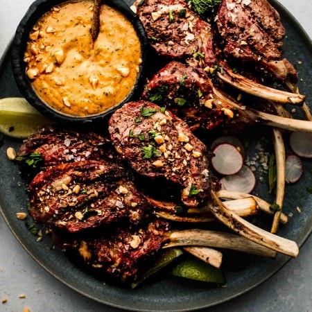 Lamb chops arranged on grey plate next to bowl of peanut sauce.