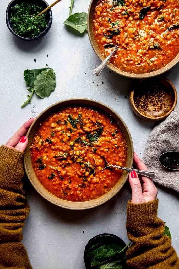 Hands holding spoon and bowl of lentil soup arranged next to bowl of red pepper flakes.
