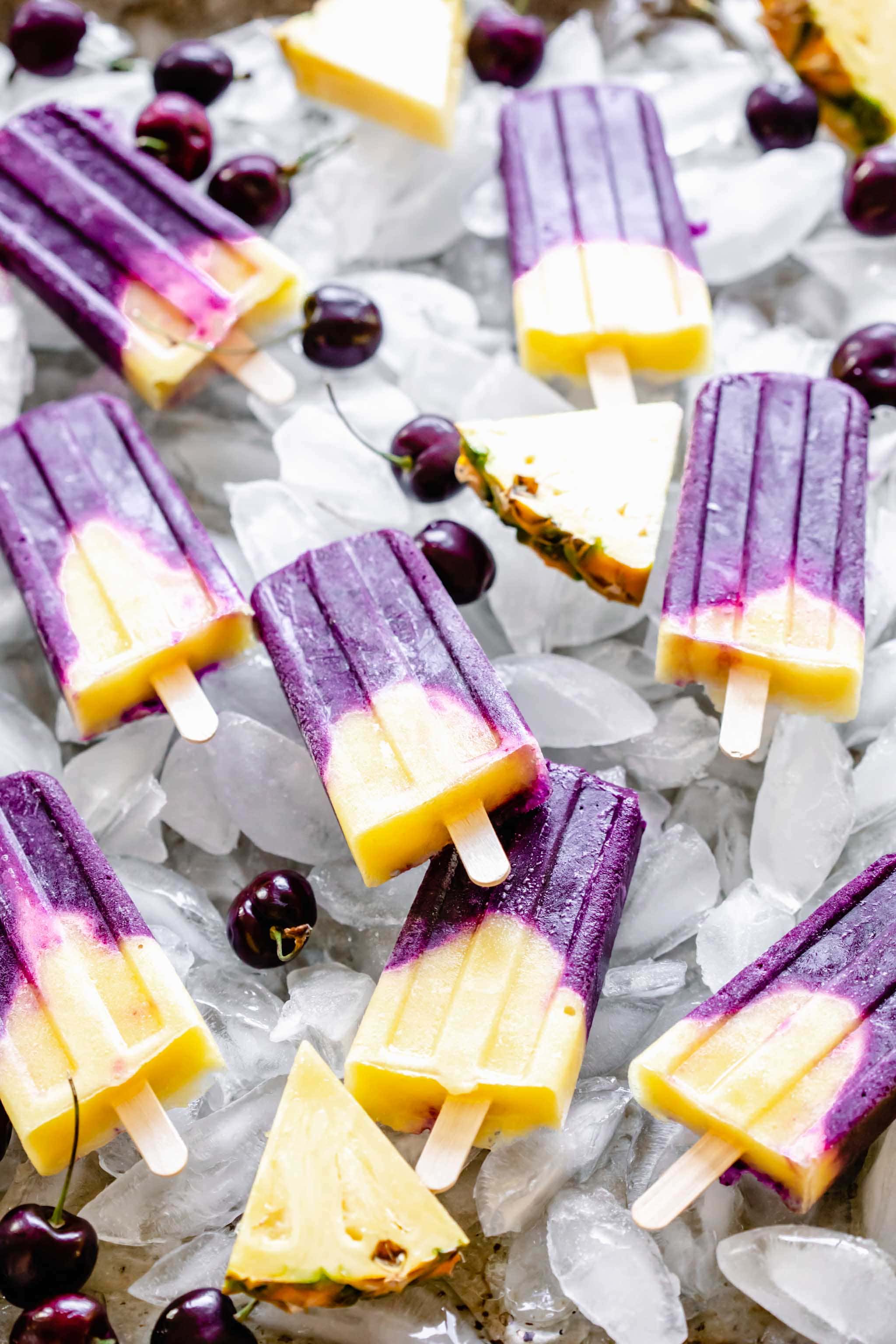 Cherry pineapple popsicles on tray of ice cubes with pineapple wedges and cherries scattered about.