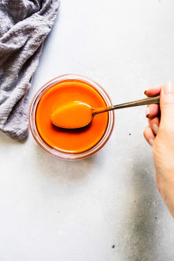 Hand holding spoon dipped into jar of buffalo sauce.