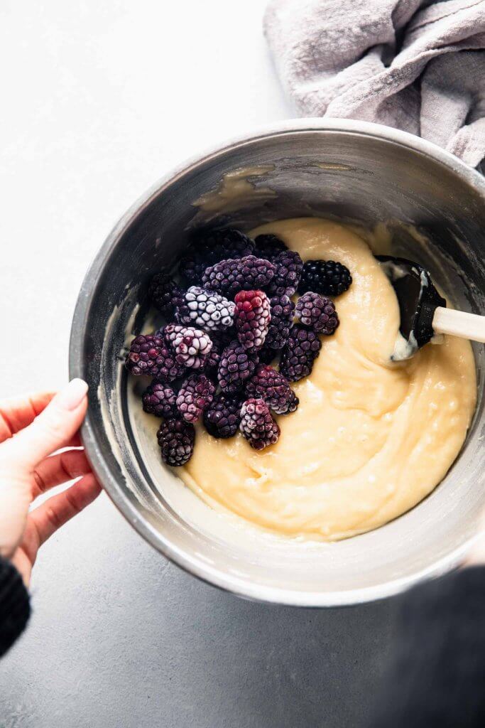 hand holding mixing bowl with mixing batter and frozen blackberries.