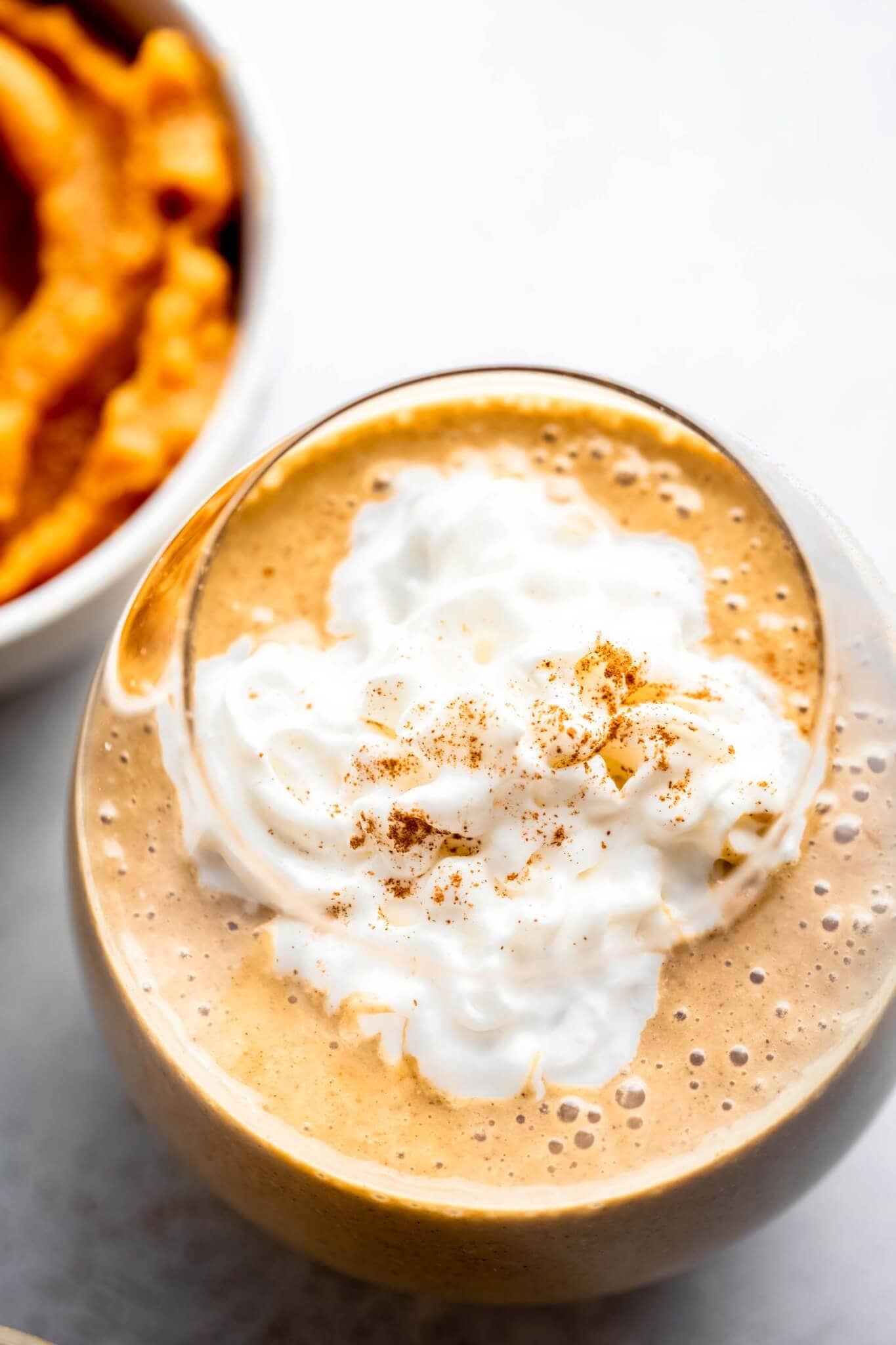 Overhead shot of pumpkin smoothie topped with whipped cream and cinnamon, next to bowl of pumpkin on light grey background.