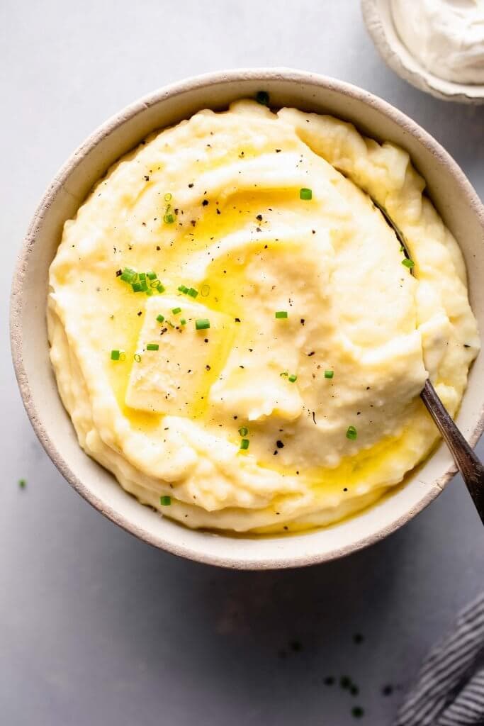 Overhead shot of bowl of mashed potatoes topped with a pat of butter, next to bowl of sour cream.