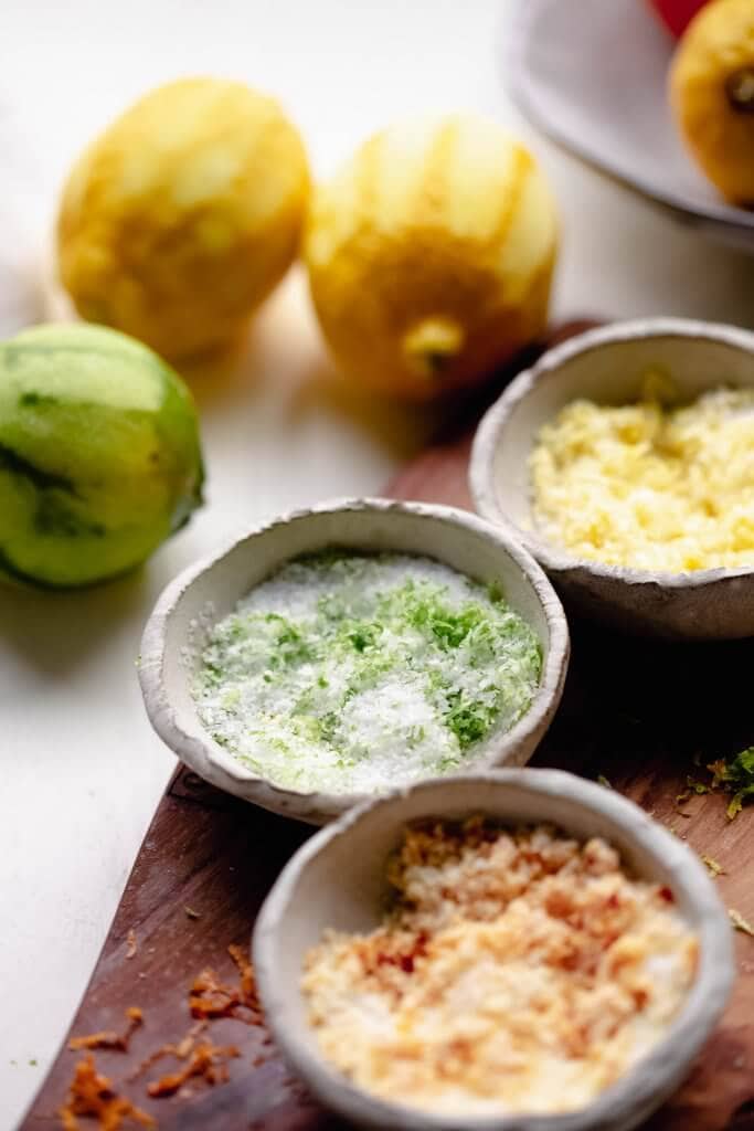 Three small bowls of lime, lemon and orange salt on cutting board next to zested citrus fruits.