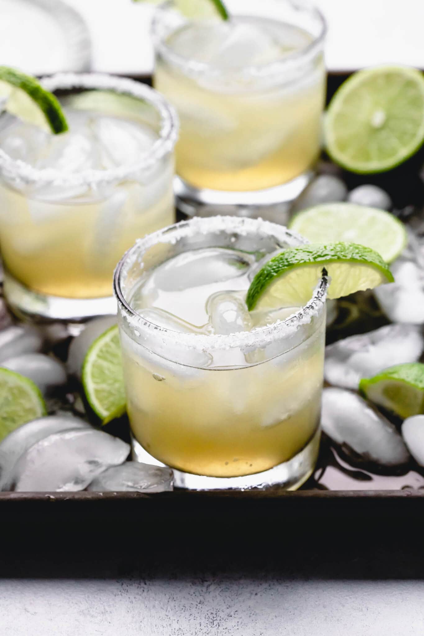 Side view of three margaritas on tray with ice cubes and limes.