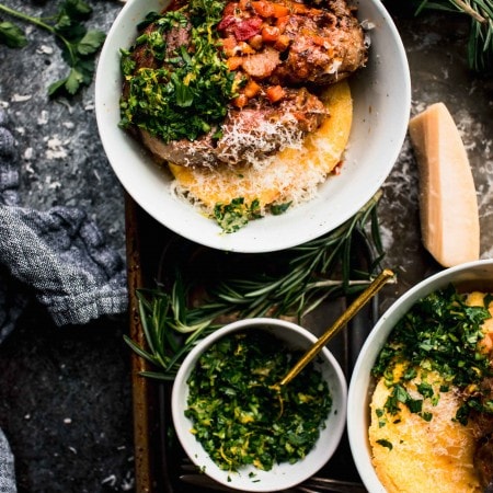 Two bowls of prepared osso buco on top of polenta on serving tray next to sprigs of fresh herbs and bowl of gremolata.