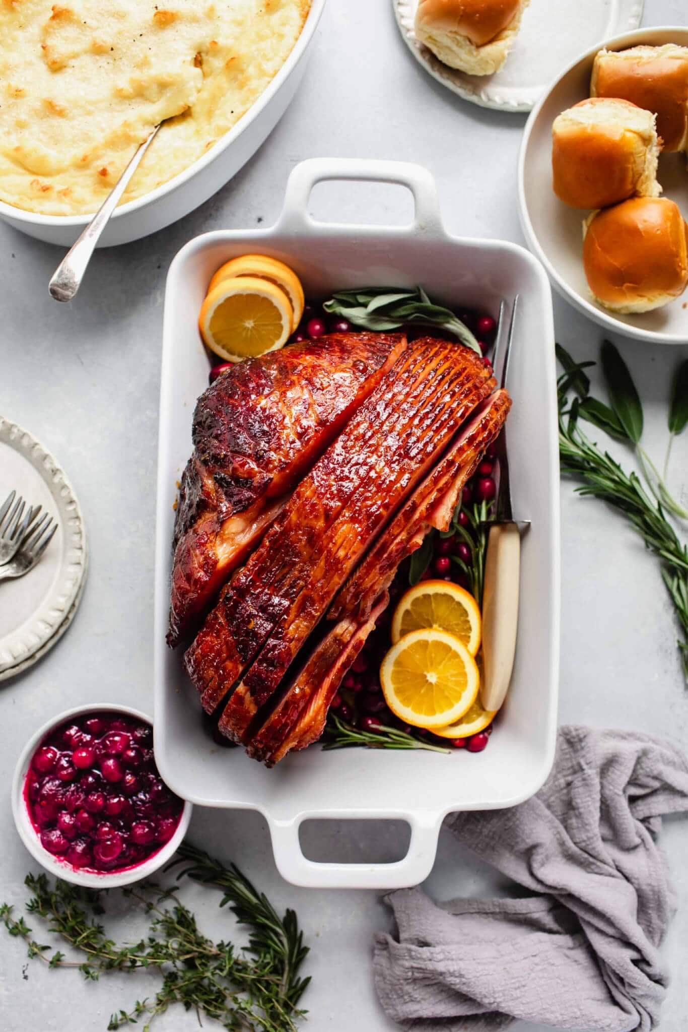 Glazed ham in roasting dish garnished with orange slices, served with mashed potatoes, cranberry sauce and rolls.