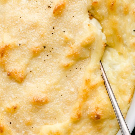 Overhead shot of baked goat cheese mashed potatoes in white casserole dish with serving spoon.