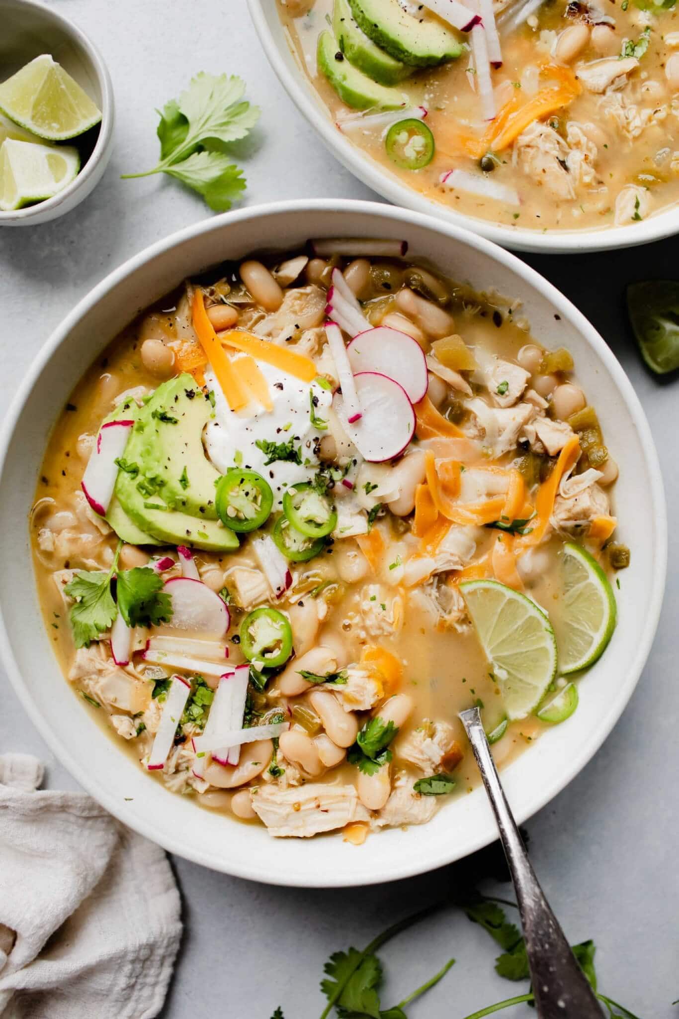 2 bowls of white chicken chili topped with cheese, sour cream and radishes.