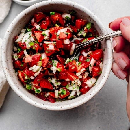 Hand dipping spoon into bowl of pico de gallo.