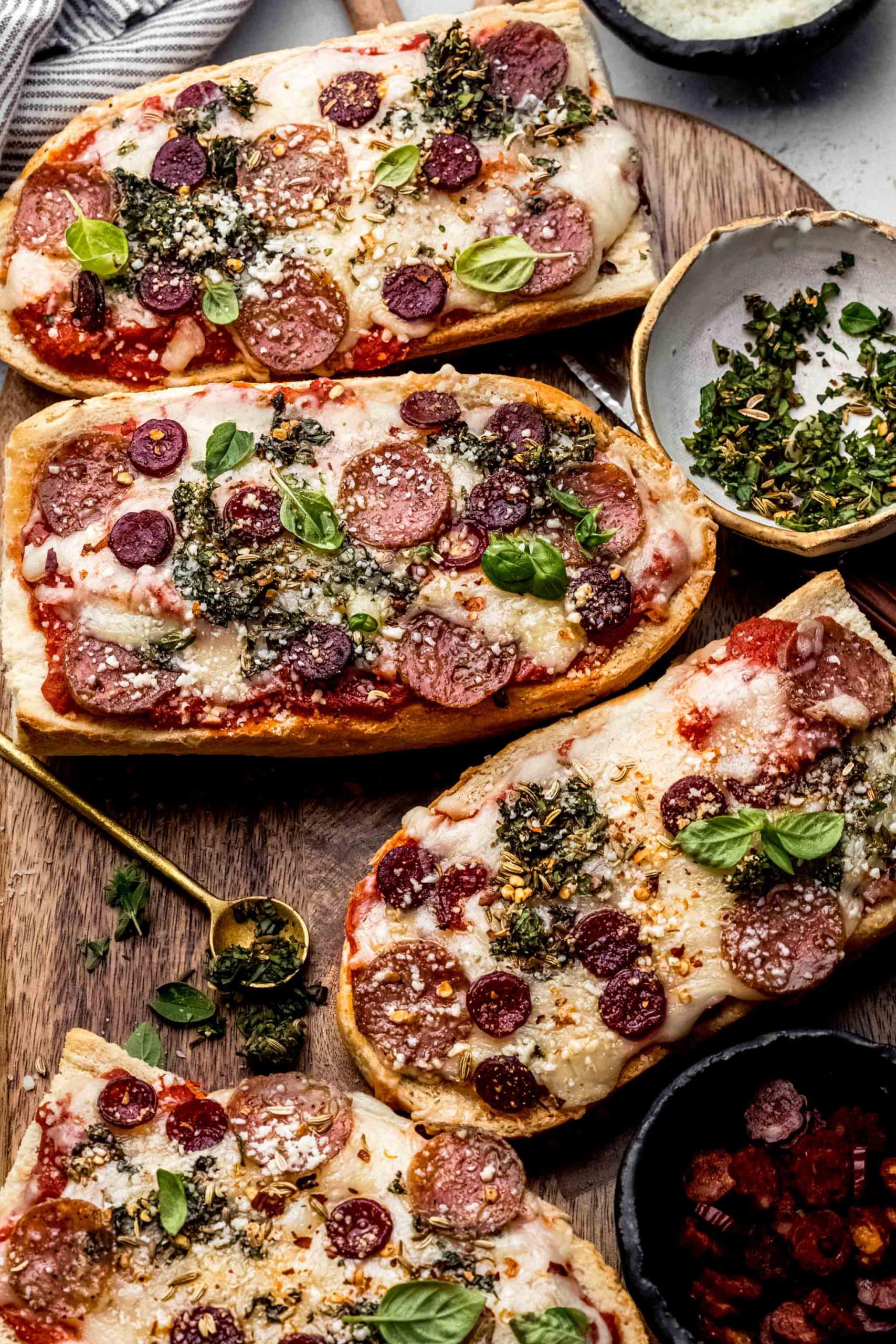 French bread pizzas on cutting board next to small bowl of herbs and pepperoni.