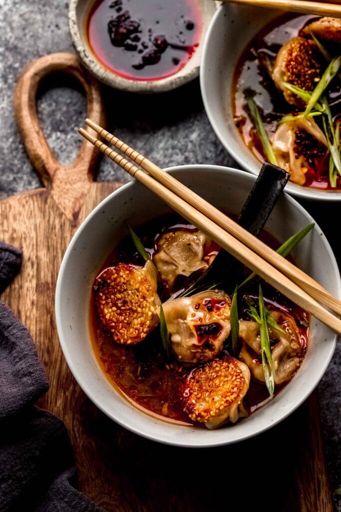 LAMB DUMPLINGS IN BOWL WITH CHOPSTICKS NEXT TO SMALL BOWL OF CHILI OIL.