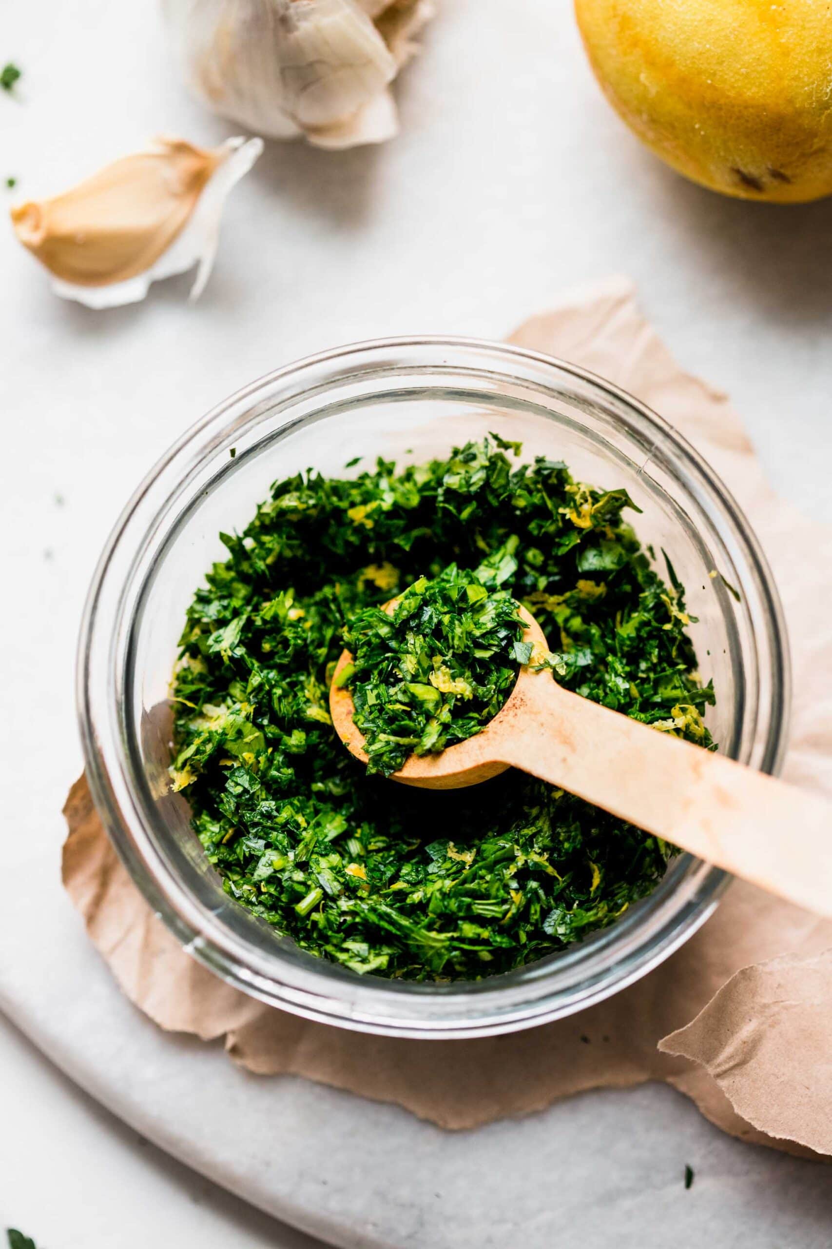 SIDE VIEW OF BOWL OF GREMOLATA NEXT TO LEMON AND GARLIC CLOVES.