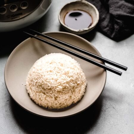 Bowl of rice with chopsticks next to rice cooker machine.