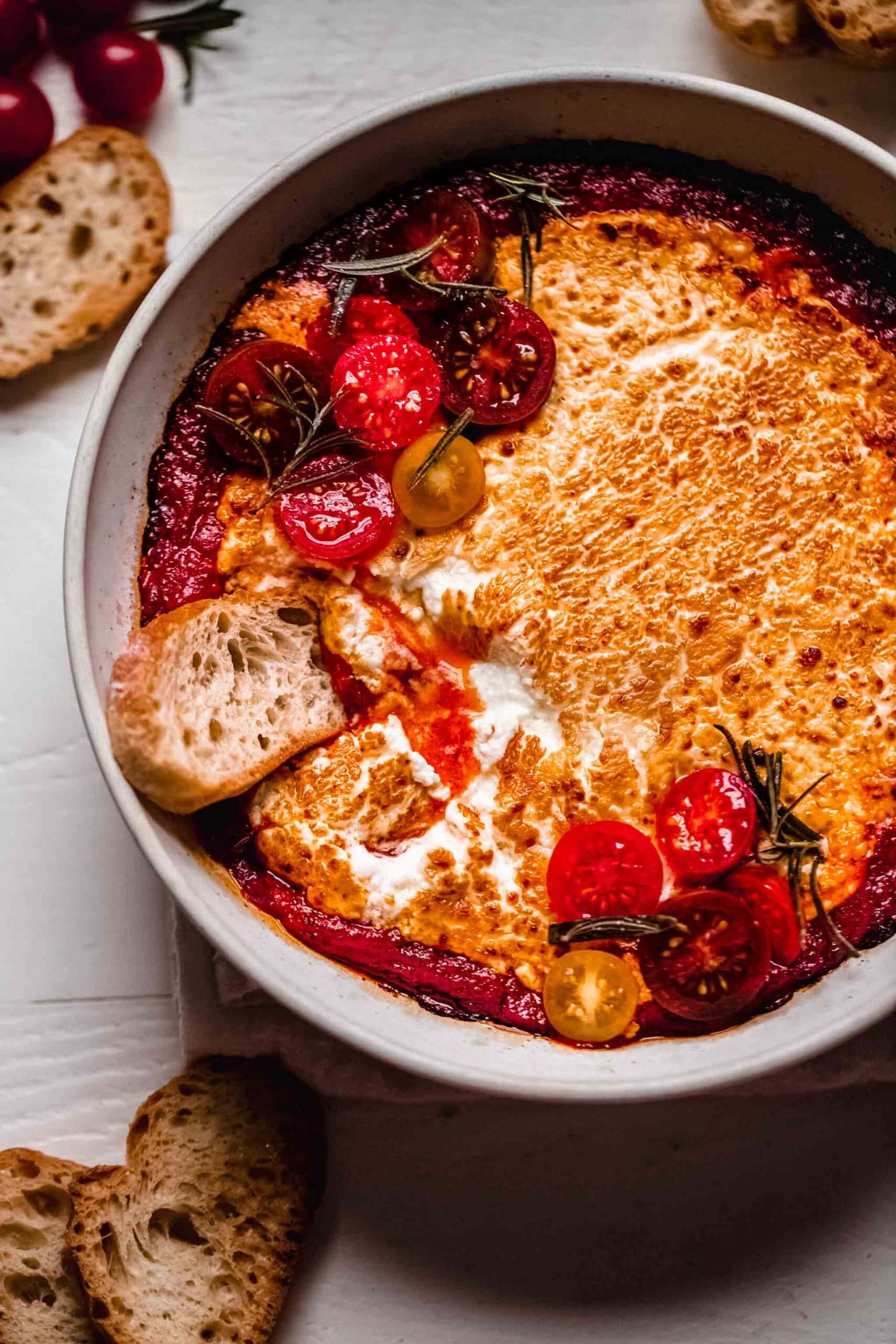 Baked goat cheese in white bowl with crostini being dipped in.