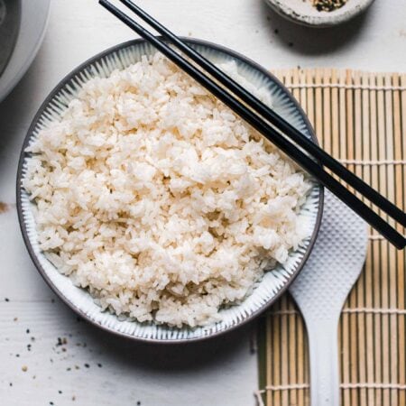 Bowl of sushi rice next to rice cooker and sushi rolling mat.