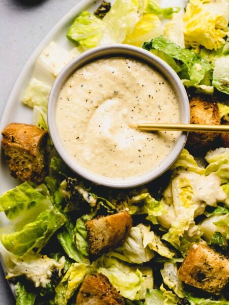 Bowl of vegan caesar dressing on plate of greens with croutons.