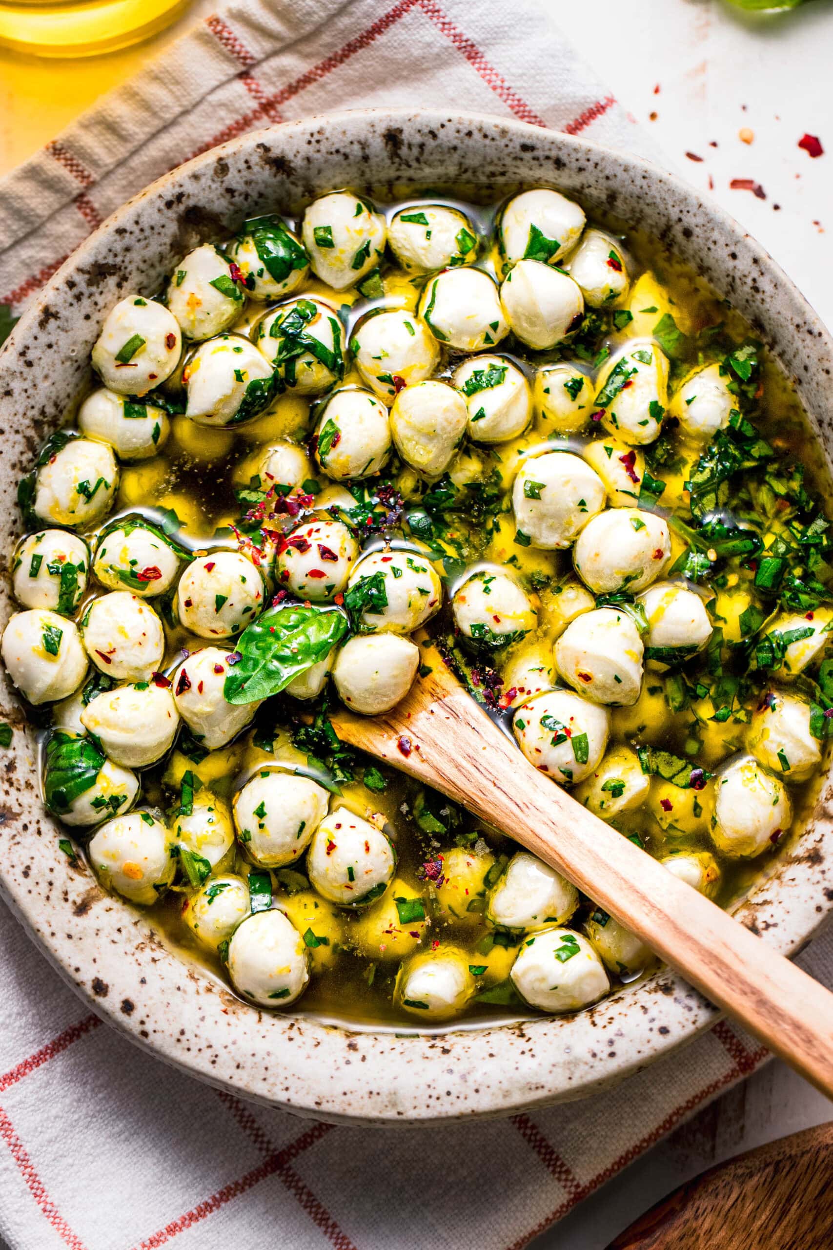Overhead shot of marinated mozzarella balls in speckled bowl with wooden spoon.
