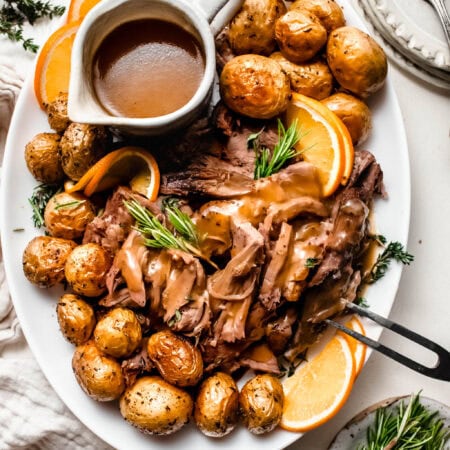 Braised lamb shoulder shredded and arranged on serving plate with orange wedges and drizzled with gravy.
