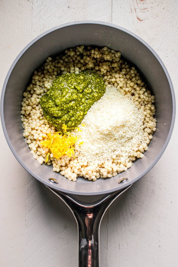 Pot of cooked israeli couscous topped with dollop of pesto, parmesan and lemon zest.