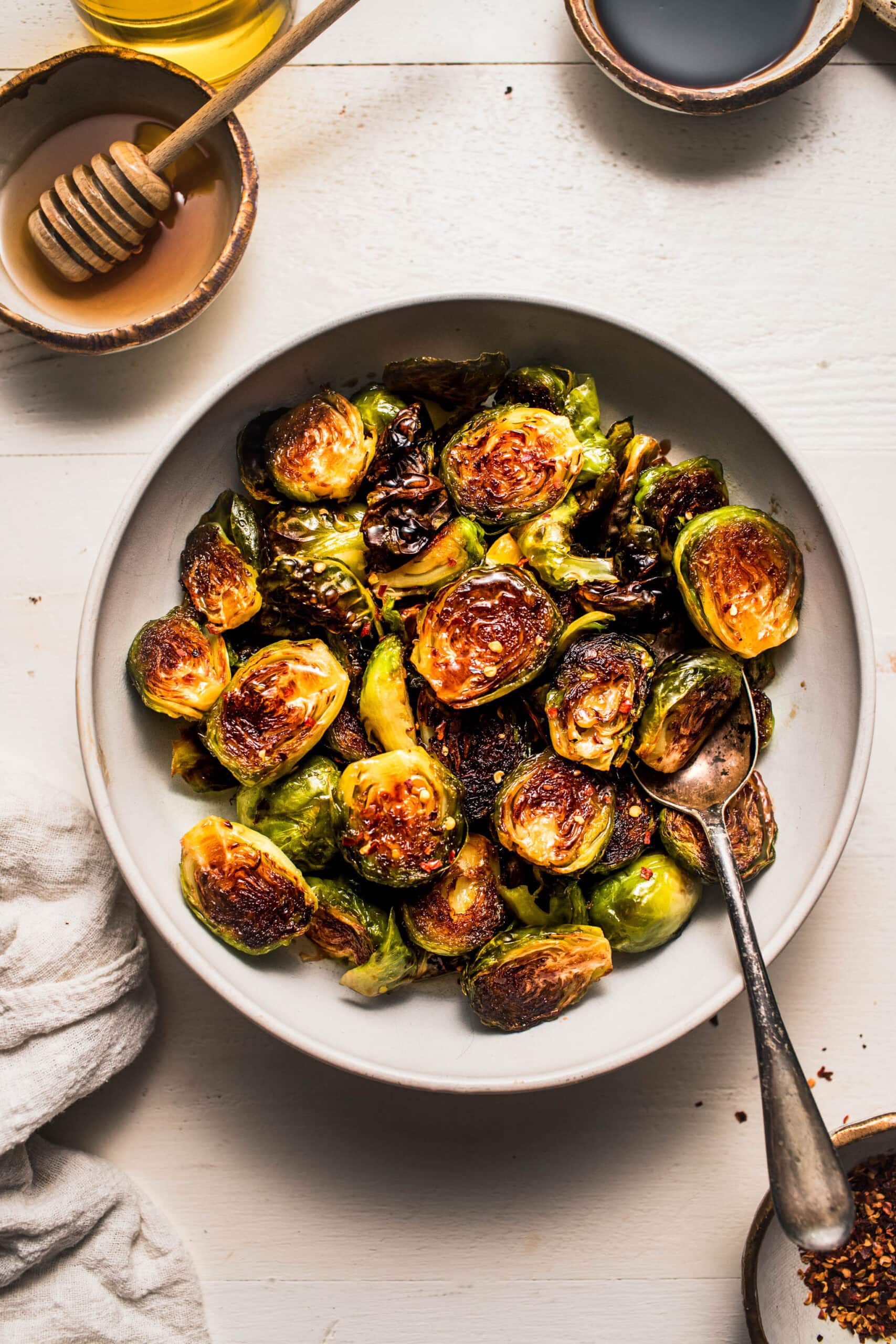 Overhead shot of honey balsamic brussels sprouts in white bowl next to bowl of honey and bowl of balsamic glaze.