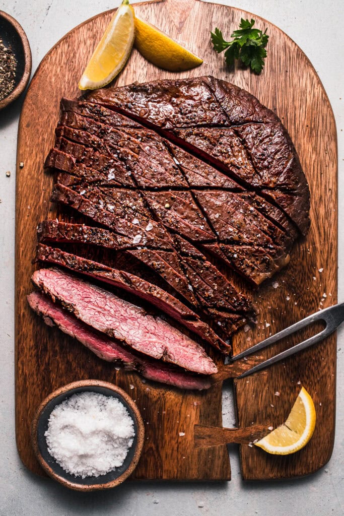 Flank steak sliced on cutting board next to small bowl of salt and lemon wedges.