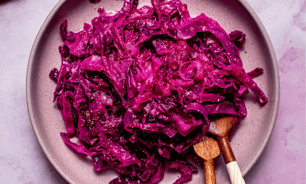 Horizontal image of Finished braised purple cabbage in pink bowl on purple counter with serving spoons.