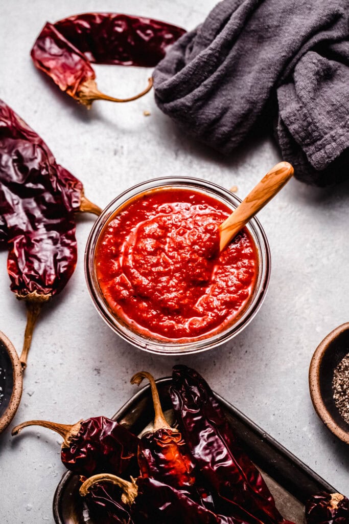 Overhead shot of finished Guajillo sauce in glass bowl with wooden spoon next to dried peppers.