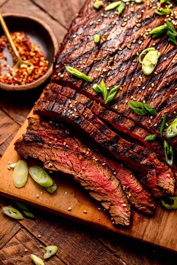 Side view of grilled flank steak on wooden cutting board sprinkled with green onions and sesame seeds.