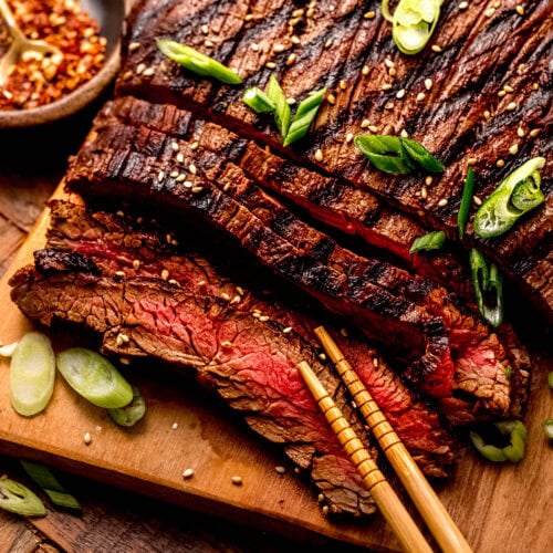 Side view of sliced and grilled asian flank steak on wooden cutting board sprinkled with green onions and sesame seeds.