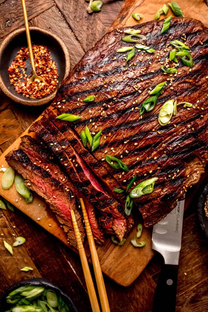 Overhead shot of flank steak with grill marks sliced on cutting board and sprinkled with green onions and sesame seeds.