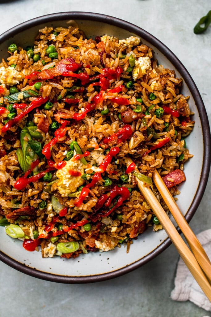 Overhead close up of fried rice in serving bowl with chopsticks drizzled with sriracha.