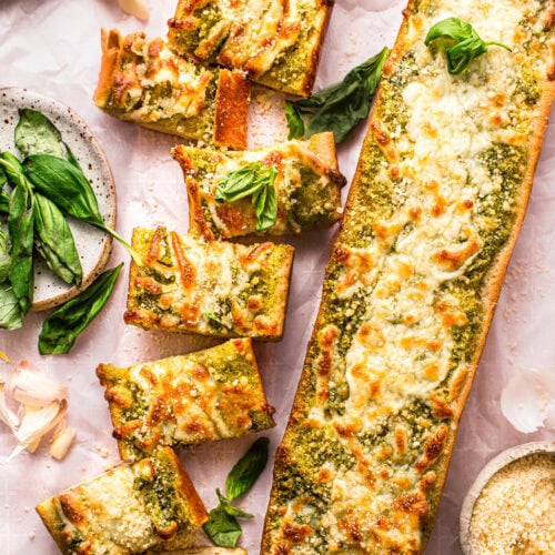 Pesto garlic bread loaves on counter being sliced into topped with basil leaves.