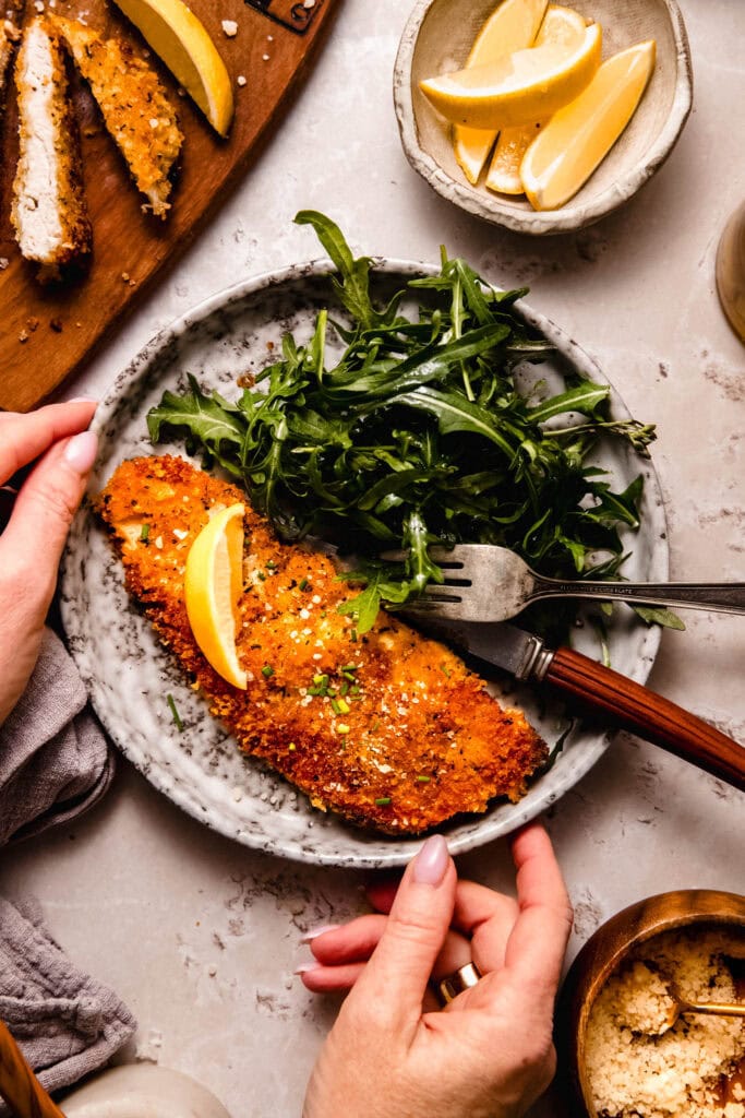 Crispy parmesan panko chicken breast cutlet on plate topped with lemon wedge and served with arugula salad.