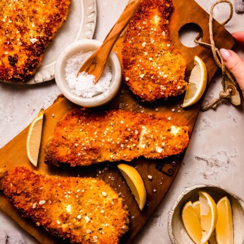 Chicken cutlets on cutting board next to bowl of flaky salt and lemon wedges.