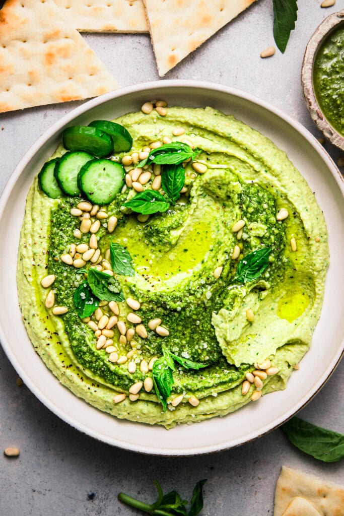 Finished pesto hummus in white bowl topped with basil sprigs and cucumber slices.