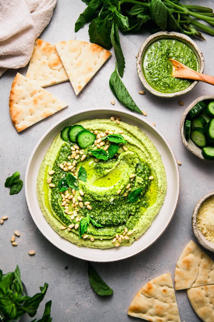 Bowl of pesto hummus on counter next to pita bread and small bowl of pesto.