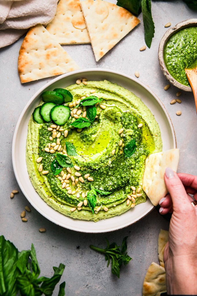 Overhead shot of piece of naan dipping into bowl of pesto hummus.