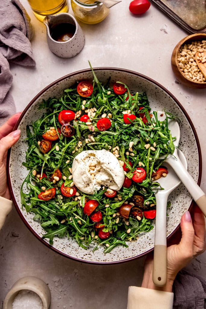 Hands holding arugula burrata salad in serving bowl.
