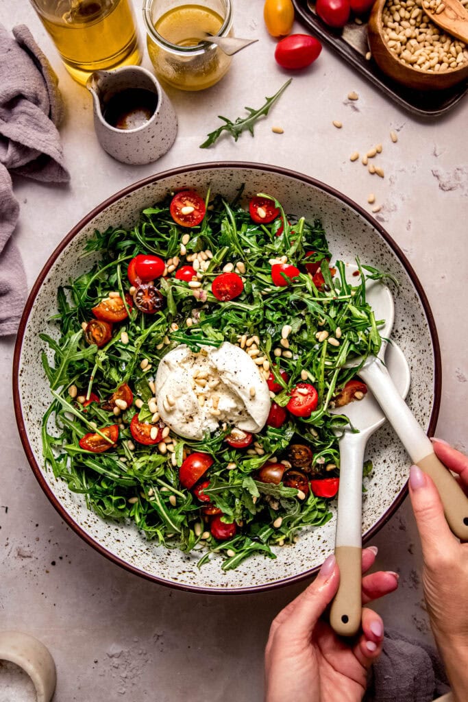 Hands holding salad tongs tossing arugula burrata salad.
