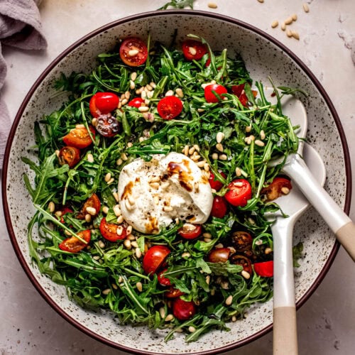 Arugula burrata salad on counter next to small jar of dressing and cherry tomatoes.
