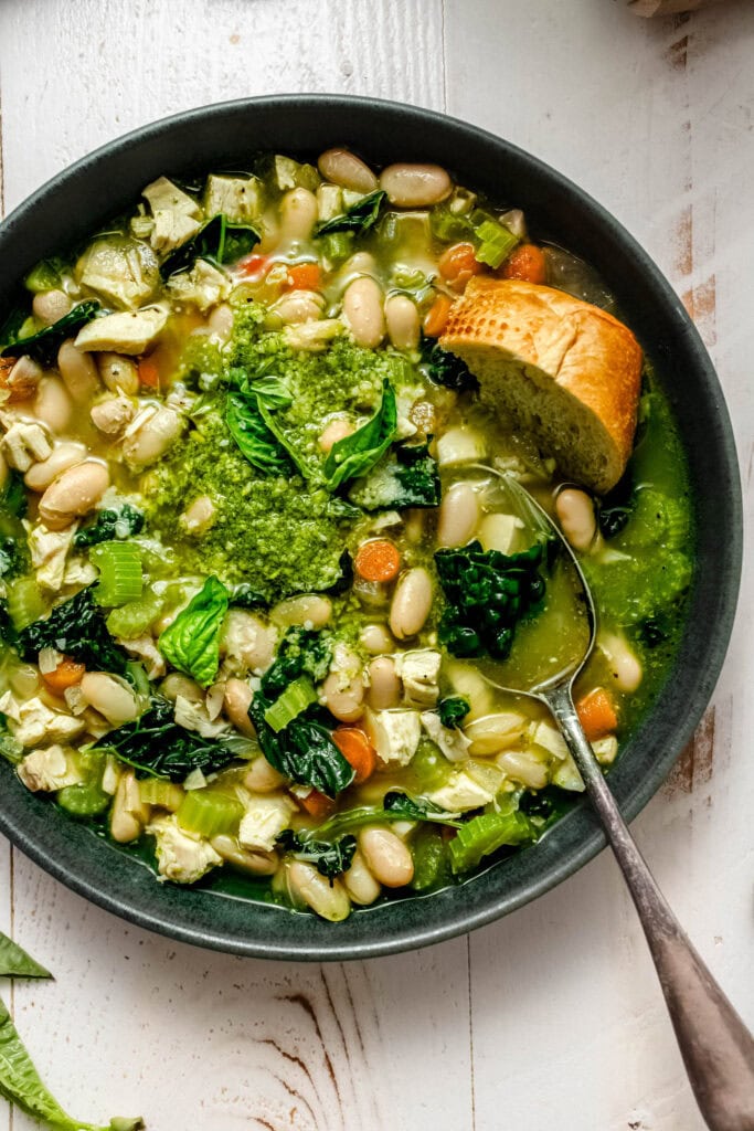 Green bowl of chicken soup with pesto, finished with a swirl of pesto and a small bowl of pesto nearby.