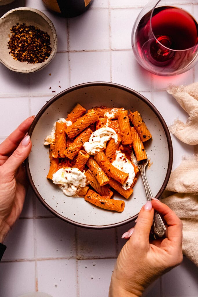 Hands holding Smoky tomato pasta with burrata cheese in serving bowl with fork.
