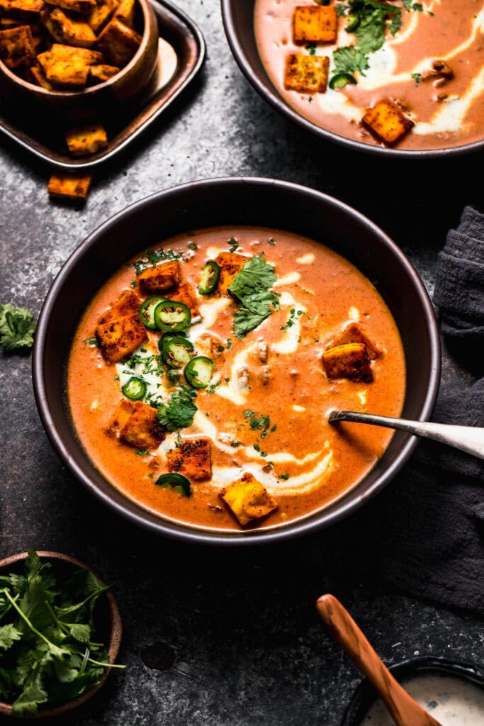 Side view of bowl of butter chicken soup in black bowl topped with naan croutons.