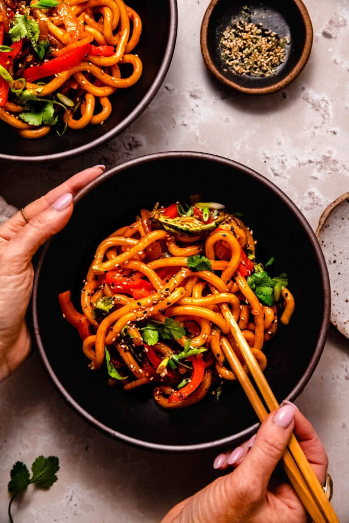 Hands holding bowl of spicy udon noodles with chopsticks.